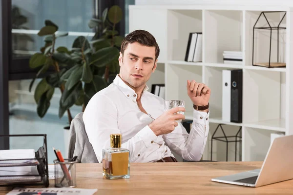 Красивый серьезный бизнесмен со стаканом виски сидит на рабочем месте в офисе — стоковое фото