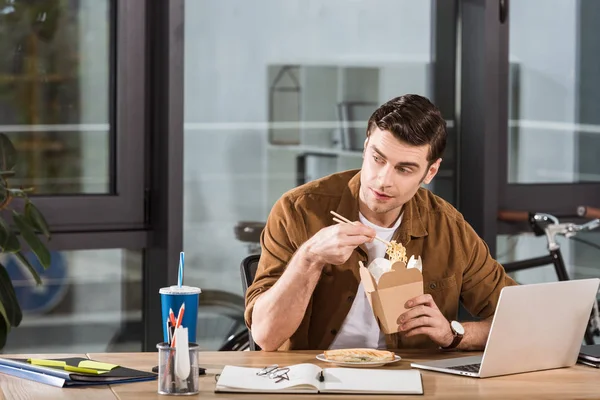 Красивый бизнесмен ест лапшу из коробки в офисе — стоковое фото