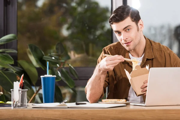 Beau homme d'affaires mangeant emporter des nouilles de la boîte au bureau et regardant la caméra — Photo de stock