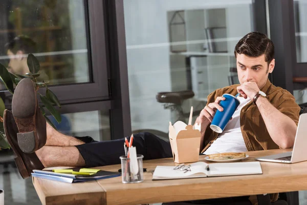 Atractivo hombre de negocios almorzando y bebiendo refrescos en el lugar de trabajo en la oficina - foto de stock