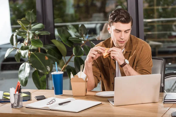 Guapo hombre de negocios comiendo sándwich y llevar fideos en la oficina mientras trabaja con el ordenador portátil - foto de stock