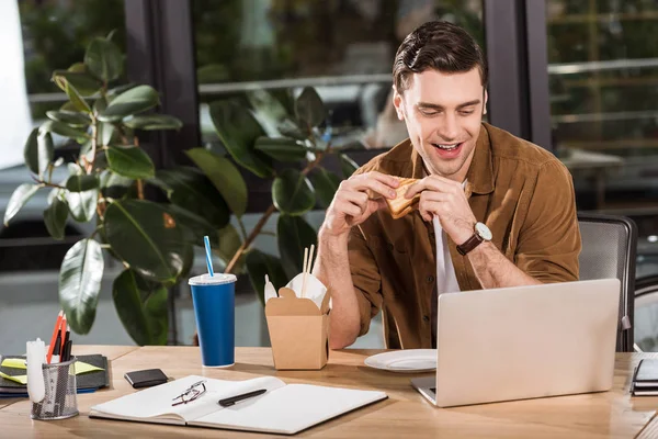 Красивый счастливый бизнесмен ест сэндвич и забирает лапшу в офисе во время работы с ноутбуком — стоковое фото