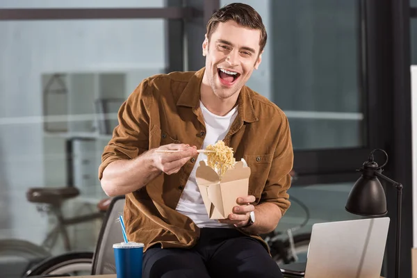 Gutaussehender, glücklicher Geschäftsmann isst im Büro Nudeln zum Mitnehmen — Stockfoto