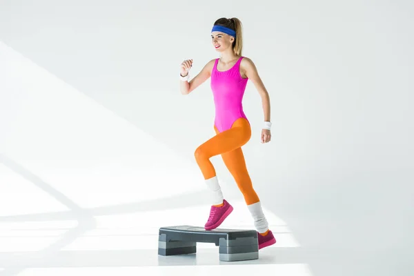Mujer deportiva sonriente ejercitando en la plataforma de paso y mirando hacia otro lado en gris - foto de stock