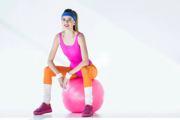 Atlética joven sentada en la pelota de fitness y sonriendo a la cámara en gris - foto de stock
