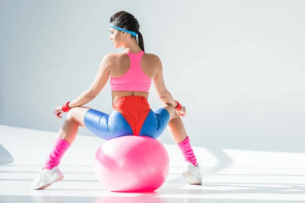 Вид ззаду на спортивну молоду жінку, яка сидить і займається фітнес-кулькою на сірому — стокове фото