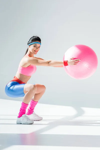 Atractiva joven entrenando con pelota de fitness y sonriendo a la cámara en gris - foto de stock