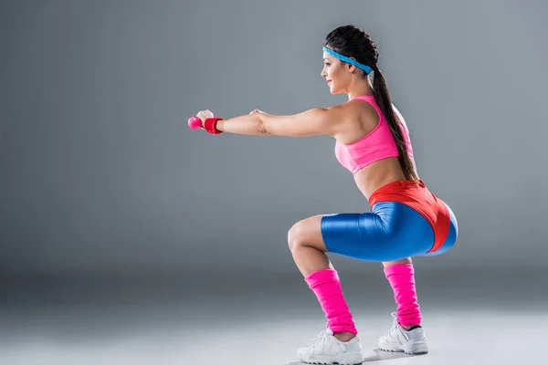 Side view of sporty girl doing squats with dumbbells on grey — Stock Photo