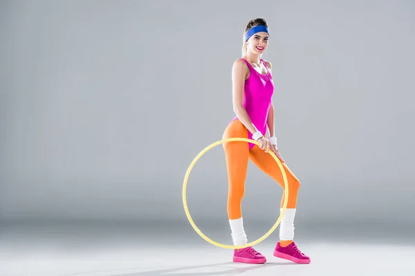 Bela menina desportiva de pé com arco de hula e sorrindo para a câmera no cinza — Fotografia de Stock