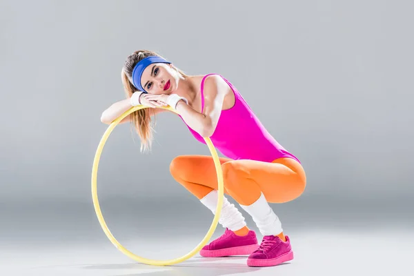 Attractive young sportswoman leaning at hula hoop and smiling at camera on grey — Stock Photo