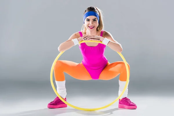 Happy young sportswoman exercising with hula hoop and smiling at camera on grey — Stock Photo