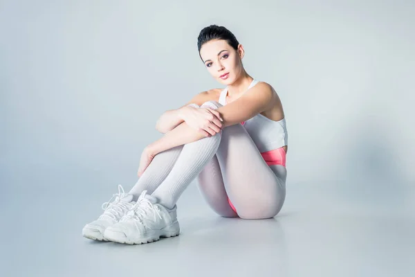 Attractive young sportswoman in bodysuit sitting and looking at camera on grey — Stock Photo