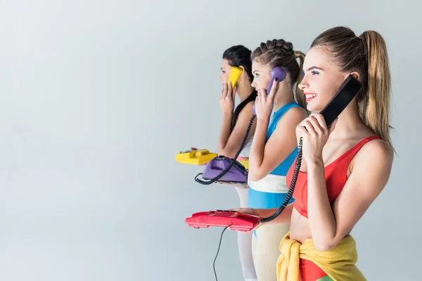 Side view of smiling sporty girls talking by rotary phones and looking away isolated on grey — Stock Photo