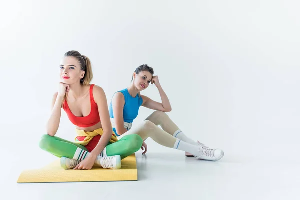 Красивые улыбающиеся спортивные девушки, сидящие на коврике для йоги и отводящие взгляд от серого — стоковое фото