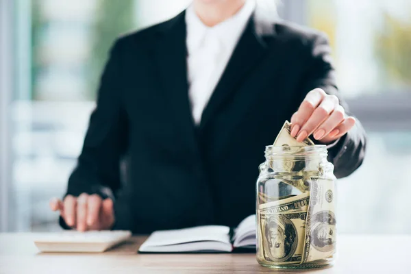 Cropped shot of businesswoman working and putting dollar banknote in glass jar — Stock Photo