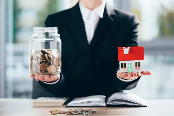 Cropped shot of businesswoman holding glass jar with coins and small house model — Stock Photo