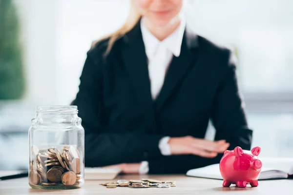 Vista close-up do banco piggy rosa e frasco de vidro com moedas, mulher de negócios sorridente atrás — Fotografia de Stock