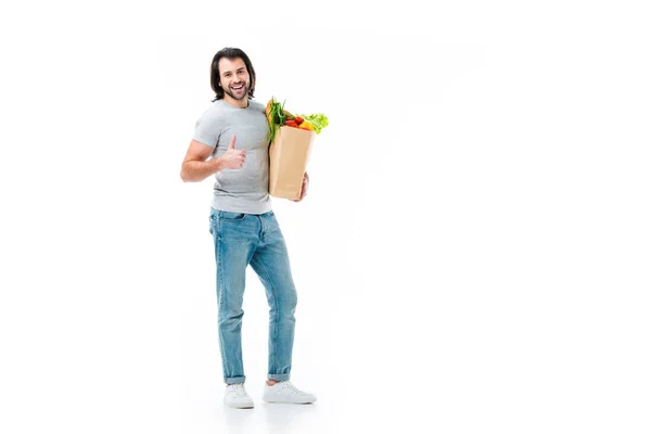 Handsome man keeping grocery bag,  showing thumb up and smiling at camera isolated on white — Stock Photo