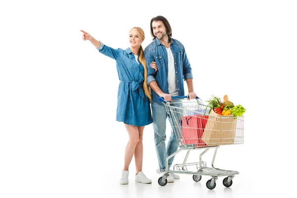 Couple with shopping cart full of bags pointing at something isolated on white — Stock Photo