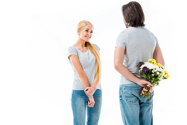 Preciosa pareja mirándose el uno al otro, hombre sosteniendo flores detrás de la espalda aisladas en blanco - foto de stock