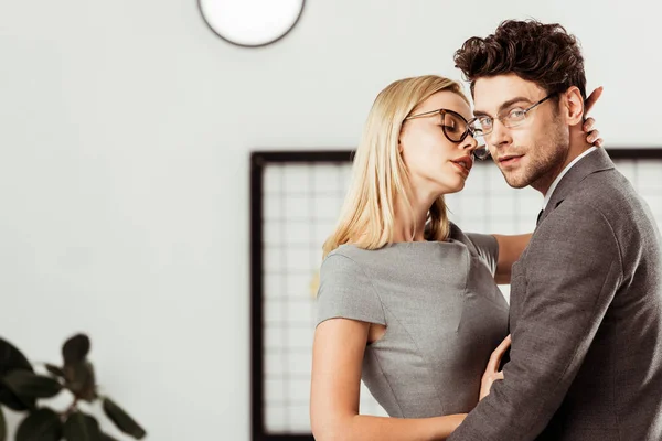 Vista lateral de la mujer de negocios abrazando colega que mirando a la cámara en la oficina, concepto de romance de oficina - foto de stock