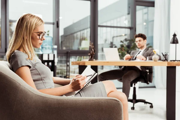 Бізнес-леді робить нотатки в блокноті, сидячи в кріслі з колегою на робочому місці в офісі — стокове фото