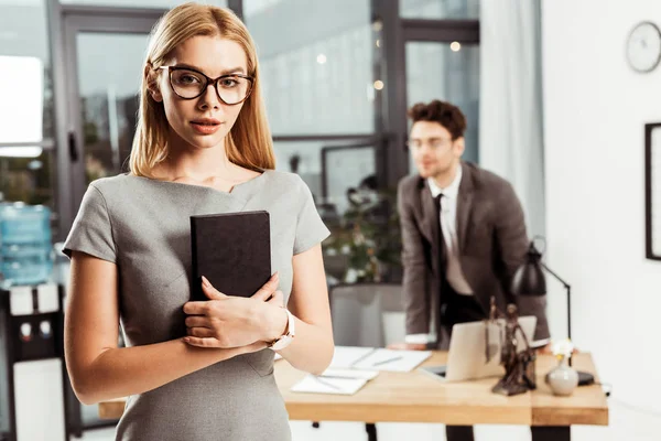 Selektiver Fokus einer jungen Rechtsanwältin mit Notizbuch, die in die Kamera blickt, während die Kollegin im Büro am Arbeitsplatz steht — Stockfoto