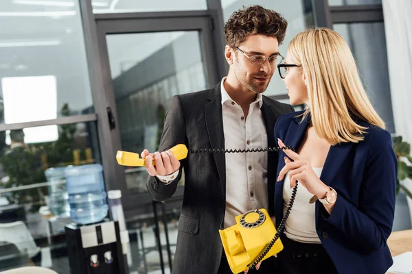 Hombre de negocios coqueteando con joven colega con teléfono retro amarillo en la oficina - foto de stock