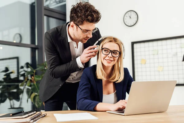 Porträt eines Geschäftsmannes, der mit einem Kollegen in Kopfhörern am Arbeitsplatz mit Laptop im Büro flirtet — Stockfoto