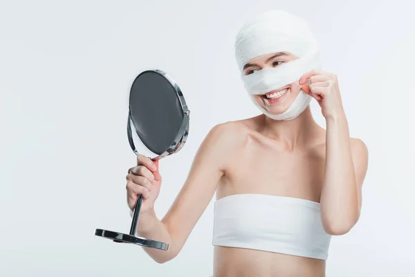 Mujer sonriente con vendajes mirando el espejo aislado en blanco - foto de stock