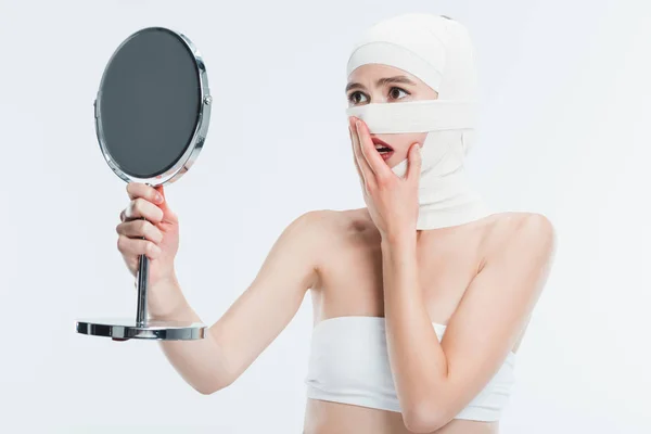 Mujer conmocionada con vendajes sobre la cabeza mirando el espejo aislado en blanco - foto de stock