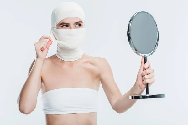 Donna con bende dopo chirurgia plastica sul viso guardando specchio isolato su bianco — Foto stock