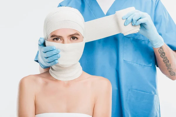 Primer plano del cirujano plástico en guantes que tapan la cara de la mujer con vendaje aislado en blanco - foto de stock