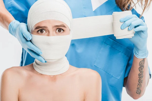 Nahaufnahme eines plastischen Chirurgen mit Handschuhen, der das verängstigte Gesicht der Frau mit einem Verband isoliert auf weißem Grund abklebt — Stockfoto