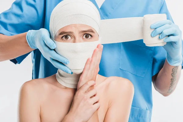 Gros plan du chirurgien plasticien en gants scotchant femme effrayée visage avec bandage isolé sur blanc — Photo de stock