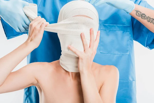Крупным пластмассовым хирургом в перчатках, заклеивающим лицо женщины повязкой, изолированной на белом — стоковое фото