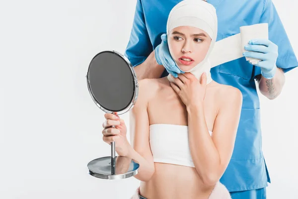 Chirurg in Handschuhen verklebt Gesicht mit Verband nach Schönheitsoperation, während Frau Spiegel isoliert auf Weiß betrachtet — Stockfoto