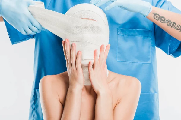 Primer plano del cirujano plástico en guantes que tapan la cara de la mujer con vendaje aislado en blanco - foto de stock