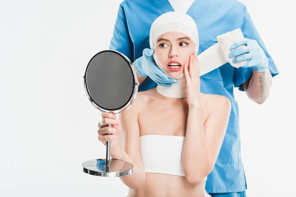 Chirurgien en gants scotcher le visage avec bandage après la chirurgie plastique tandis que la femme effrayée regardant miroir isolé sur blanc — Photo de stock