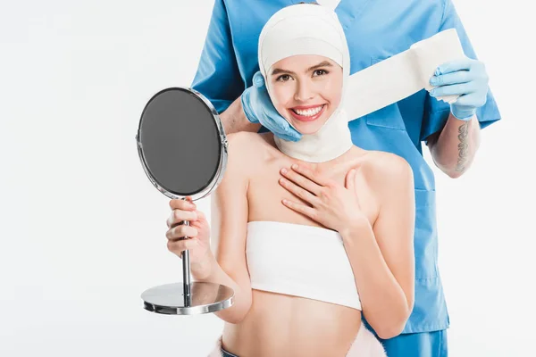 Chirurgien en gants scotcher le visage avec un bandage après une chirurgie plastique tout en souriant femme tenant miroir isolé sur blanc — Photo de stock