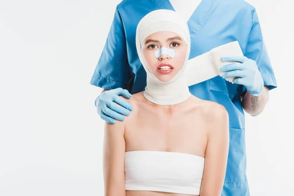 Cirurgião em luvas amarrando o rosto da mulher com bandagem após cirurgia plástica isolada em branco — Fotografia de Stock
