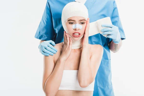 Cirurgião em luvas amarrando o rosto da mulher com bandagem após cirurgia plástica isolada em branco — Fotografia de Stock