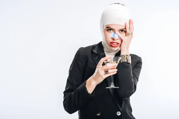 Femme élégante avec des bandages sur la tête après la chirurgie plastique tenant verre avec cocktail isolé sur blanc — Photo de stock