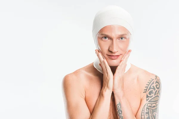 Hombre guapo adulto con vendajes sobre la cabeza tocando la cara después de la cirugía plástica aislado en blanco - foto de stock