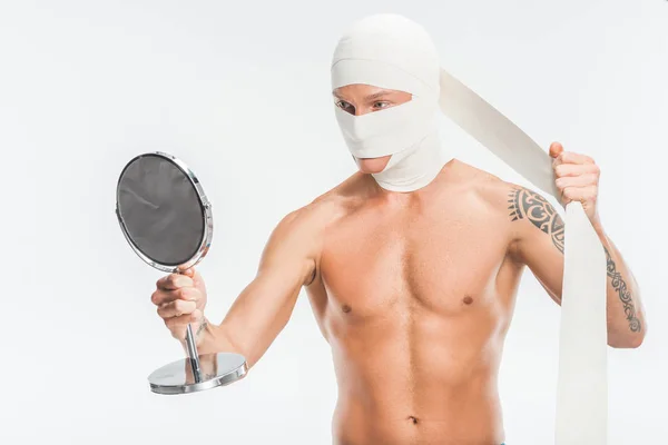 Голый мужчина снимает повязки с головы после пластической операции и смотрит на зеркало, изолированное на белом — стоковое фото