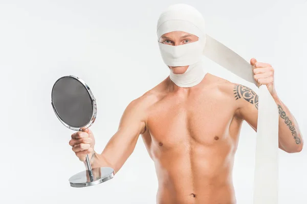 Голый мужчина снимает бинты с головы после пластической операции и держит зеркало изолированным на белом — стоковое фото