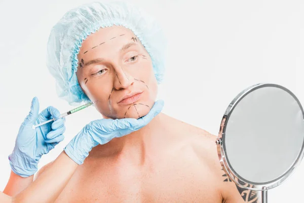 Manos del cirujano sosteniendo la jeringa mientras el hombre mira el espejo aislado en blanco - foto de stock