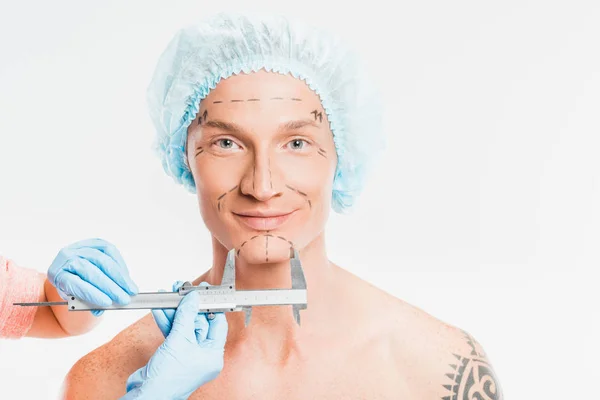 Manos médicas que miden la cara del hombre con marcas dibujadas aisladas en blanco - foto de stock