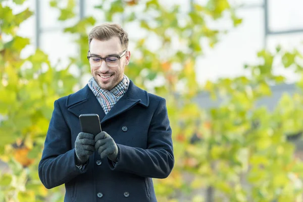 Sorrindo homem digitando no smartphone com árvore borrada — Fotografia de Stock