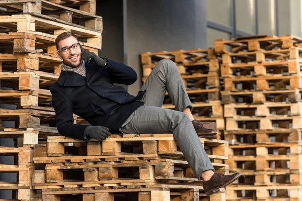 Schöner lächelnder Mann, der mit dem Smartphone spricht und auf Holzpaletten liegt — Stockfoto